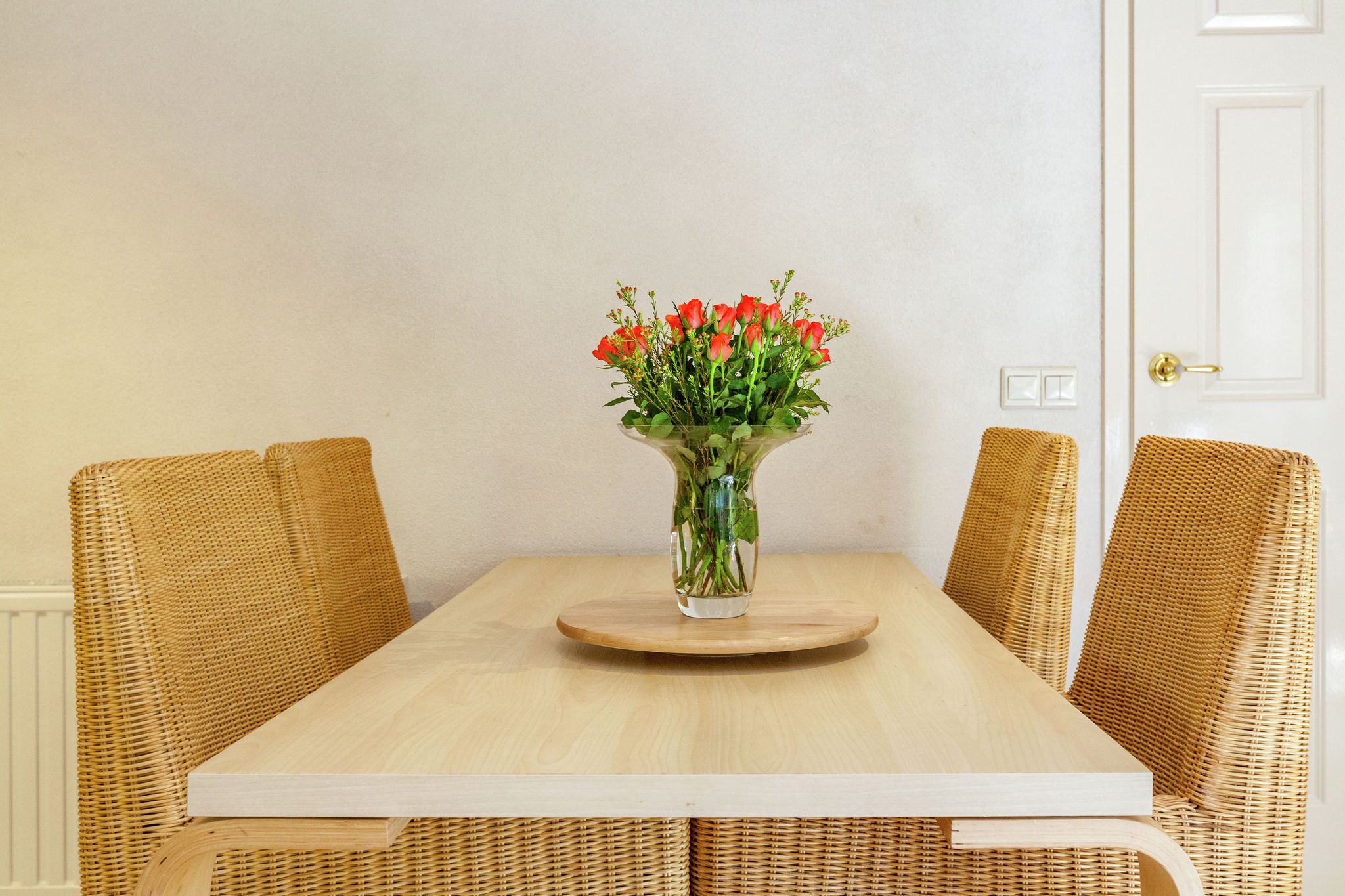 Une table à manger ornée d’un vase de fleurs rouges, entourée de chaises en osier.