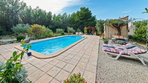 Piscina com deck revestido, cercada por vegetação e uma pérgola com área de estar ao ar livre.