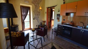 A cozy kitchen with a stone wall, wooden beams, and a rustic stove. There is a dining area with a table and chairs, and a kitchen counter with a kettle. - Countryside cottage in Pergola (Pergola)
