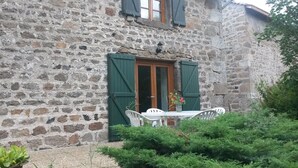 A stone building with a wooden door and green shutters, a white table with chairs, and a potted plant in the foreground. - Rustic Retreat in Goutte-Claire (Sauvain)