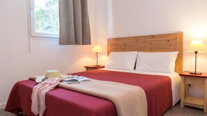 Hotellihuone, jossa on puinen sängynpääty, kaksi yöpöytää valaisimineen, punavalkoinen ja beigenvärinen sängynpeite, ikkuna verhoineen sekä pieni puinen jakkara.
