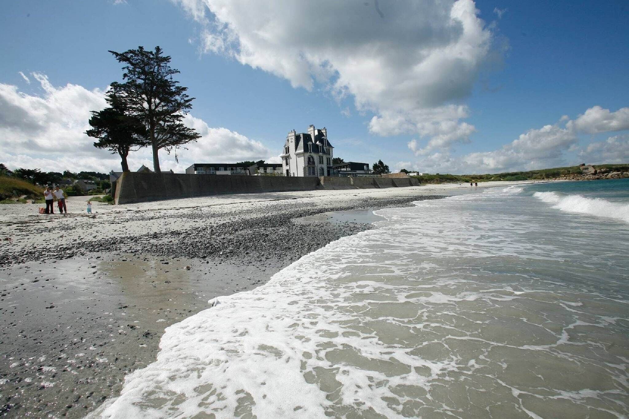 A beach with a large building in the background, a tree, and people walking along the shore.