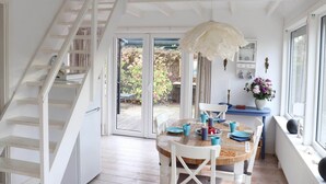 Una habitación con una mesa de comedor de madera, sillas blancas, una escalera y vistas a un jardín a través de puertas de cristal.
