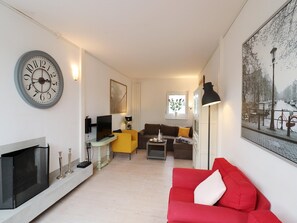 Een woonkamer met een televisie, bank en een rode slaapbank. Op de muur hangt een klok en een schilderij van een stadsgezicht.