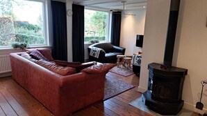 A cozy living room with a red sofa, a black armchair, a fireplace, and a large window with curtains. - Villa in Vessem near Eindhoven (Vessem)