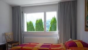 Ein Schlafzimmer mit großem Fenster, einem Sessel, einem Bett mit farbenfroher Decke und einem Heizkörper.