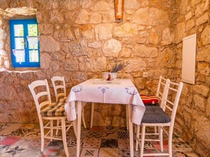 Una habitación con paredes de piedra, una mesa preparada para dos personas, dos sillas blancas, un ventanal con marco azul y un piso con diseño.