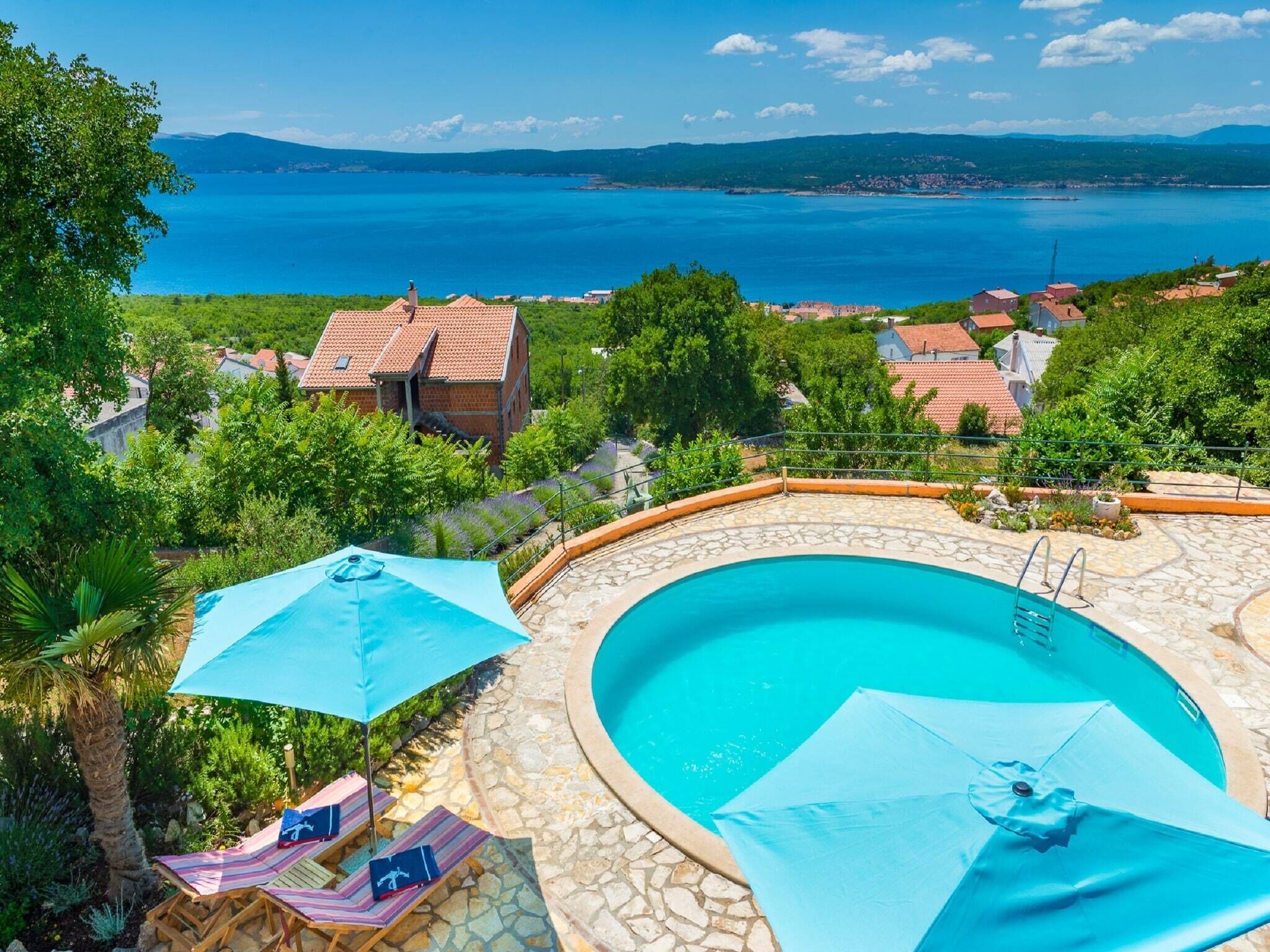 A swimming pool with a stone deck, surrounded by lounge chairs and umbrellas, overlooking a coastal landscape with houses and a clear blue sea.