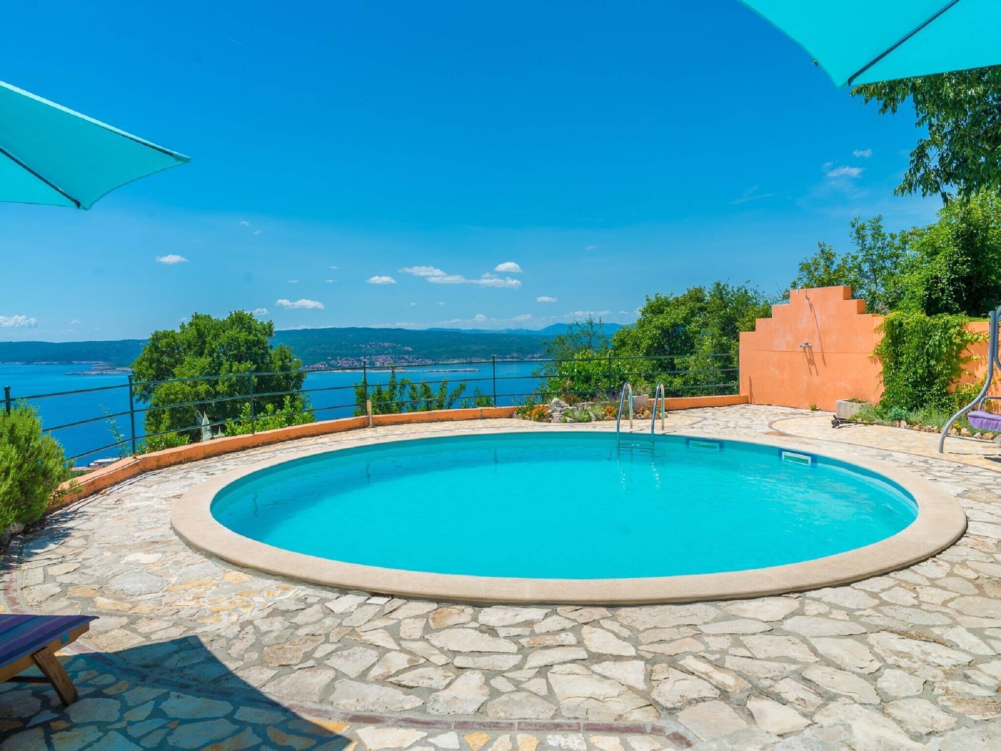 A circular pool with a stone deck, surrounded by a low wall and a view of the sea.