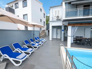 A poolside area with lounge chairs and umbrellas, a dining area with tables and chairs, and a staircase leading to an upper level.