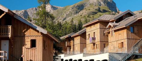 A wooden chalet with a balcony, a covered walkway, and a mountain backdrop.