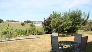 Un área de estar al aire libre con una mesa y sillas, rodeada de un jardín con diversas plantas y vistas al campo.