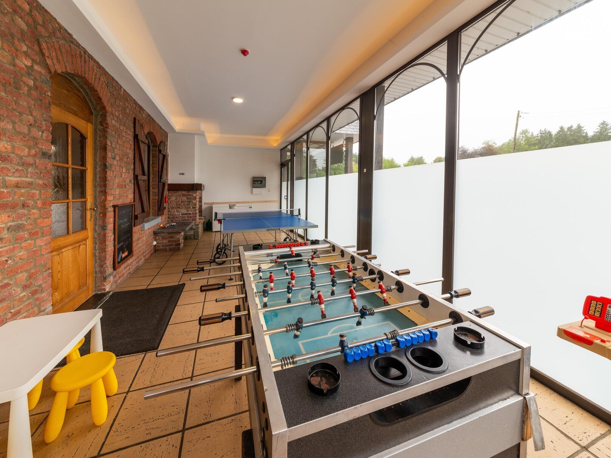 A room with a foosball table, a yellow stool, and a brick wall.