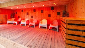 A sauna room with wooden walls, a wooden ceiling, and a wooden floor. There are six white plastic chairs arranged in two rows.