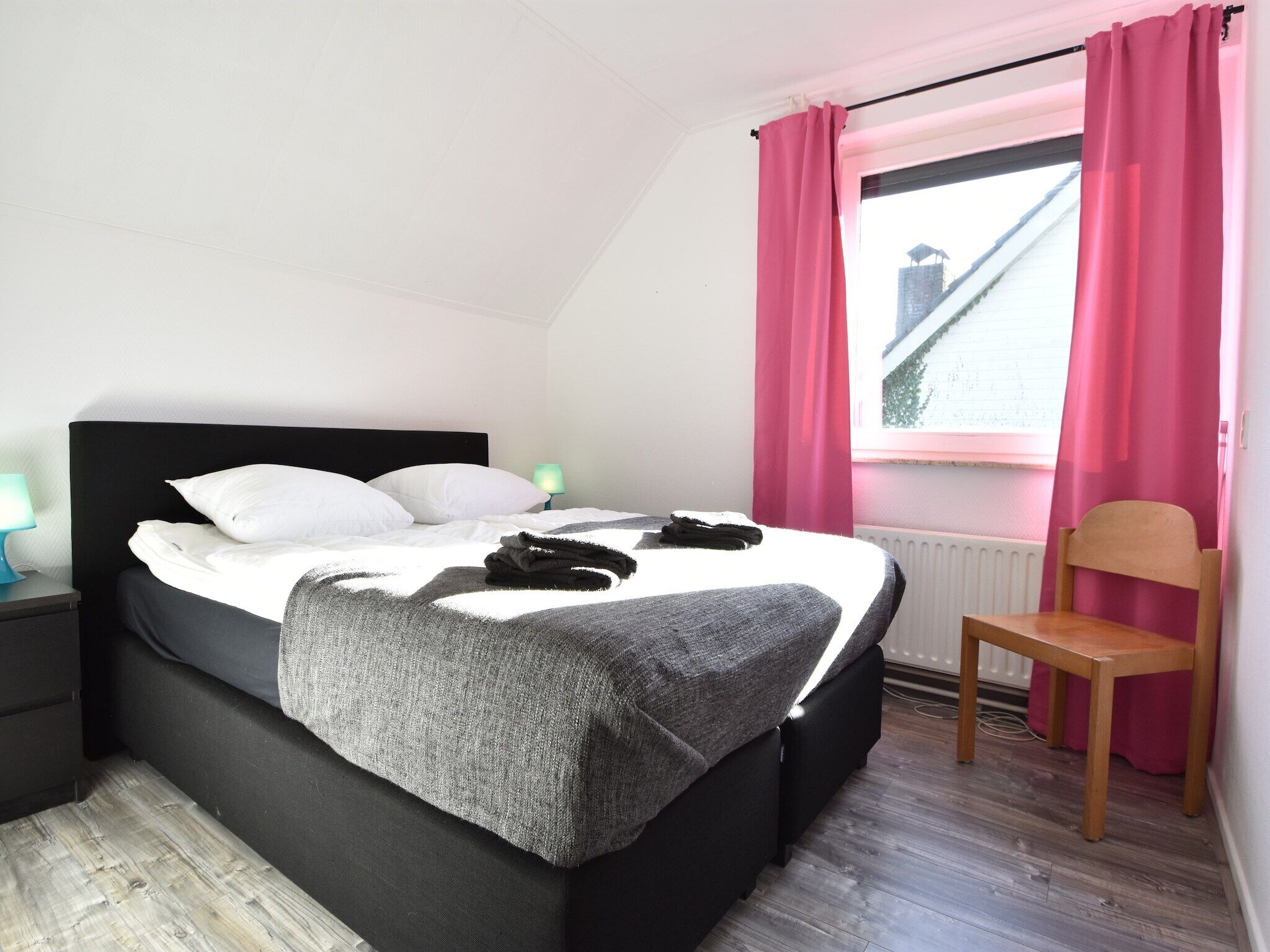 Una cama bien hecha con almohadas blancas y una manta gris, una silla de madera, una mesita de noche con una lámpara y una ventana con cortinas rosadas.