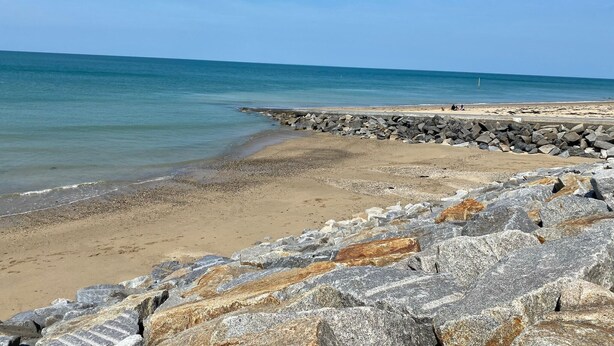 A beach with a rocky breakwater, clear blue water, and a sandy shore.