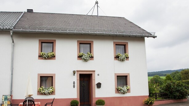 Un bâtiment à deux étages, doté d’une façade blanche et d’une base rouge. On y trouve six fenêtres ornées de jardinières, une porte sur laquelle est inscrit le numéro « 13 », et un petit jardin avec des plantes en pot.