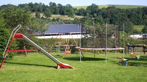 Une aire de jeux avec un toboggan, des balançoires et un trampoline, située dans un espace verdoyant, avec une maison et des arbres en arrière-plan.