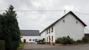 Un bâtiment à deux étages, doté d’un toit en tuiles, d’un porche à l’avant avec des chaises et d’un petit jardin agrémenté de plantes en pot.