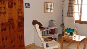 A room with a wooden wardrobe, a white chair, a small table with a plant, a bookshelf, and a floor lamp.