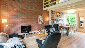 A spacious living area with a brick wall, a loft bedroom, a dining area, and a bookshelf filled with books.