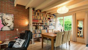 A cozy living room with a fireplace, a bookshelf, a dining area, and a large window offering a view of greenery.