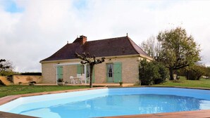 A swimming pool with a wooden deck, a stone house with green shutters, and a small patio with outdoor furniture.