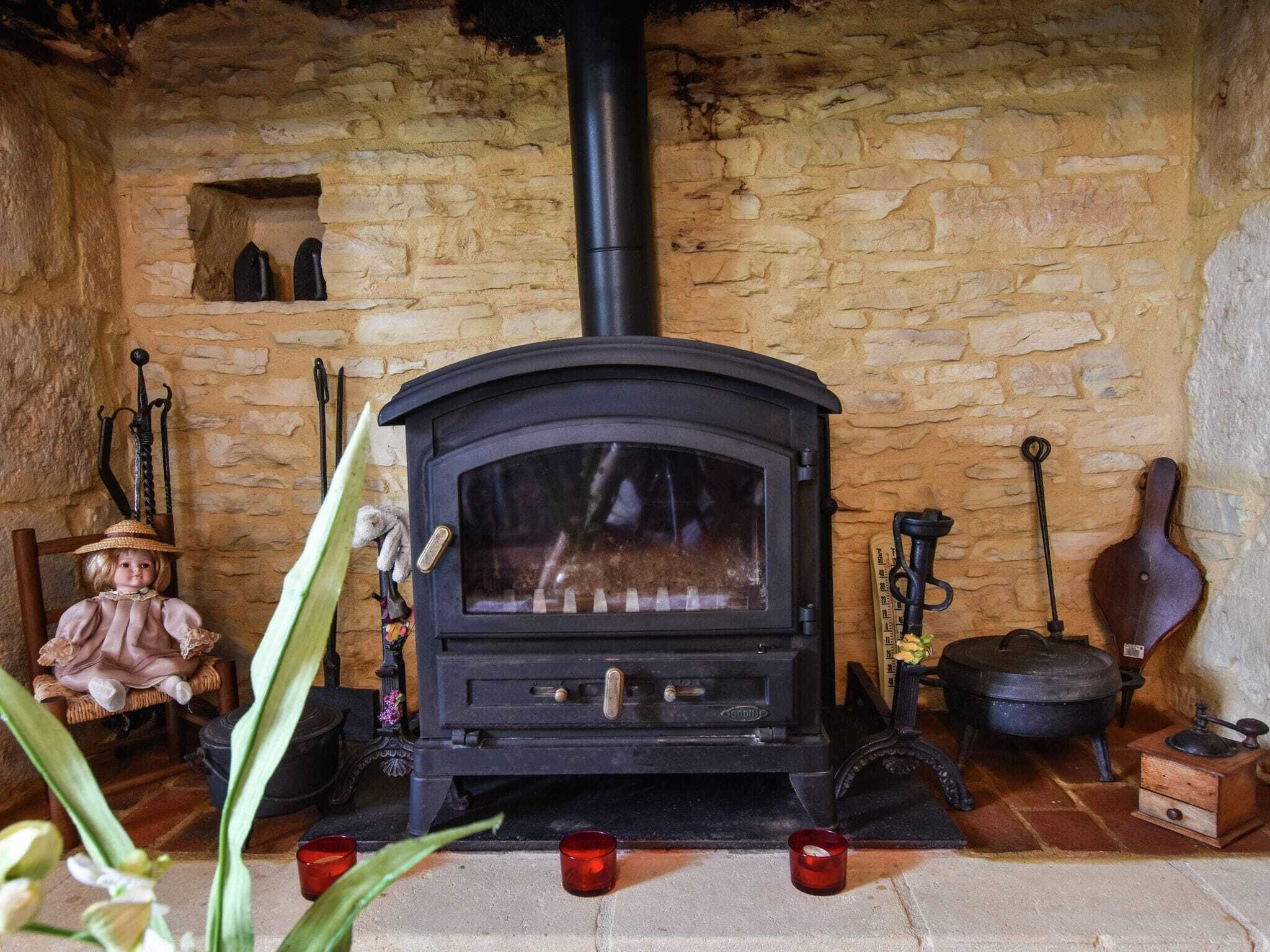 A stone-walled room with a black wood-burning stove, a small shelf with decorative items, and a rustic wooden chair.