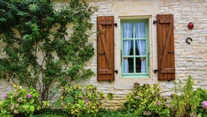 A stone building with a window and shutters, surrounded by greenery and flowers.