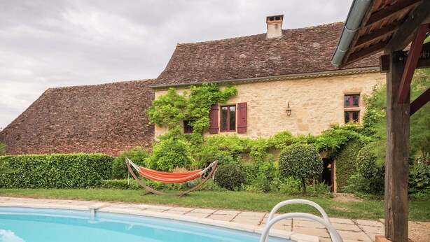 Ein Pool mit Holzterrasse, einer Hängematte und einem Steinhaus mit roten Fensterläden.