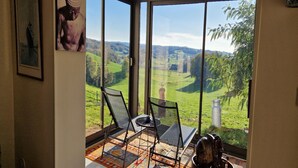 Een kamer met grote ramen met uitzicht op een groen landschap. In het midden staat een stoel en een tafel.