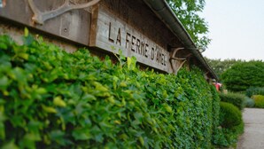 Een houten bord in rustieke stijl met de tekst 'LA FERME D'AMEL', gedeeltelijk verborgen door een haag.