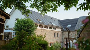 Een stenen gebouw met een schuin dak, een houten pergola en een tuin met zichtbare planten en bloemen.