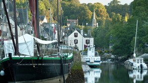 Un port où sont amarrés des bateaux, un bâtiment blanc aux volets rouges et, au loin, le clocher d’une église.