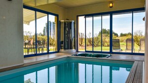 Une piscine intérieure avec des parois en verre transparent, une terrasse en bois et une vue sur l’extérieur.