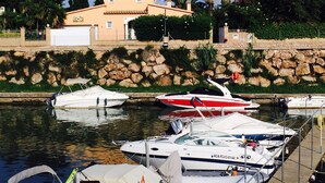Un puerto deportivo con varias embarcaciones amarradas, un muro de piedra y un edificio con techo de tejas al fondo.