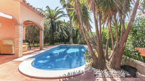 Una piscina con un borde de baldosas, palmeras y una estructura de ladrillo con una silla.