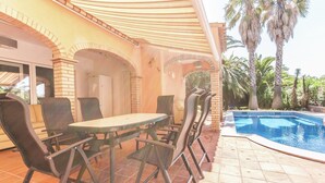 Un patio exterior con piscina, rodeado de palmeras y una zona de estar cubierta con sillas y una mesa.