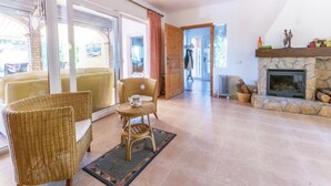 Amplio salón con chimenea de piedra, mobiliario de mimbre y vistas a través de grandes puertas corredizas de cristal.