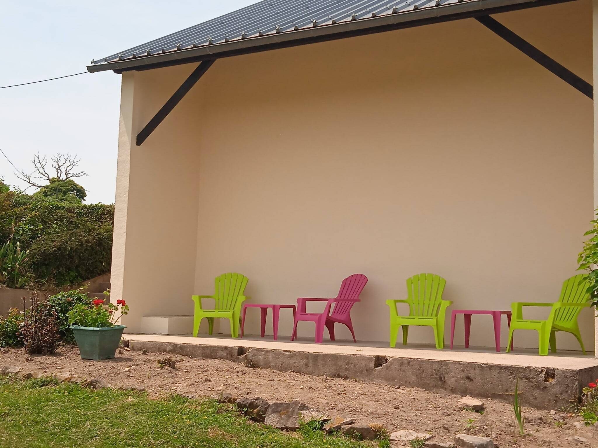 Un área de estar al aire libre con techo, con cuatro sillas y una mesa, rodeada por un muro de concreto bajo y un jardín con plantas y una maceta.