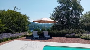 A swimming pool with two lounge chairs and an umbrella, surrounded by a landscaped garden and a view of hills. - Vacation Home in France with Pool (Vaux)