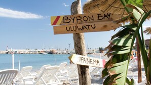A beach with white lounge chairs and wooden signs indicating Byron Bay and Ipanema.