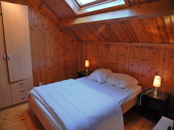 A bedroom with a skylight, wooden walls, a bed with white bedding, two bedside lamps, and a wooden wardrobe.