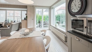 A modern kitchen with a wooden table, white chairs, a large window, and a clock on the wall. - Nice holiday home in Westende-Bad with sauna and pingpong (Westende)