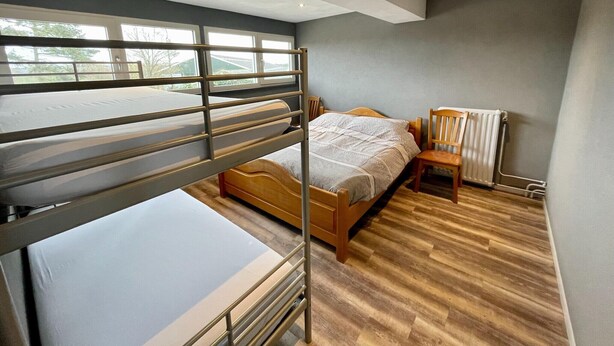 A bunk bed with a wooden frame and metal railings, a wooden chair, a radiator, and a window with a view of trees.