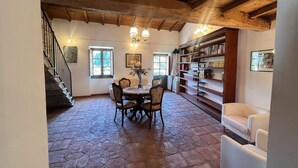 A dining area with a stone floor, a round table, and chairs. There is a staircase on the left, a bookshelf on the right, and a window with curtains.