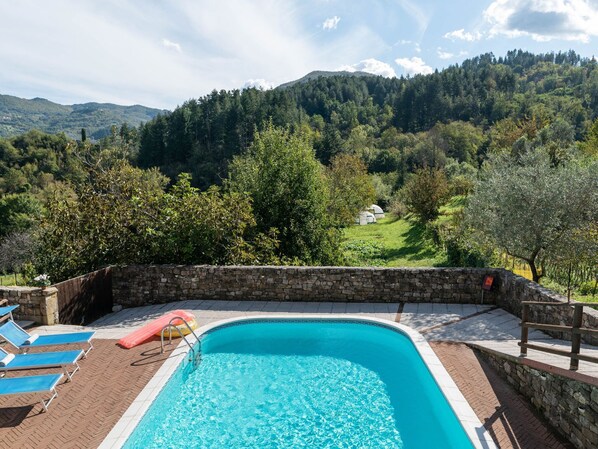 Una piscina con muro de piedra, sillas de descanso y vistas a un paisaje montañoso.