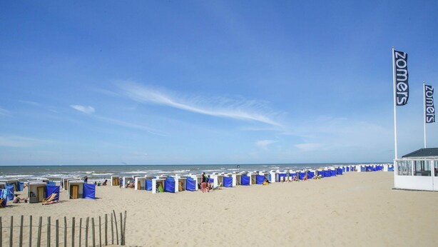 A beach with rows of blue beach huts and a flag reading "Zomers".