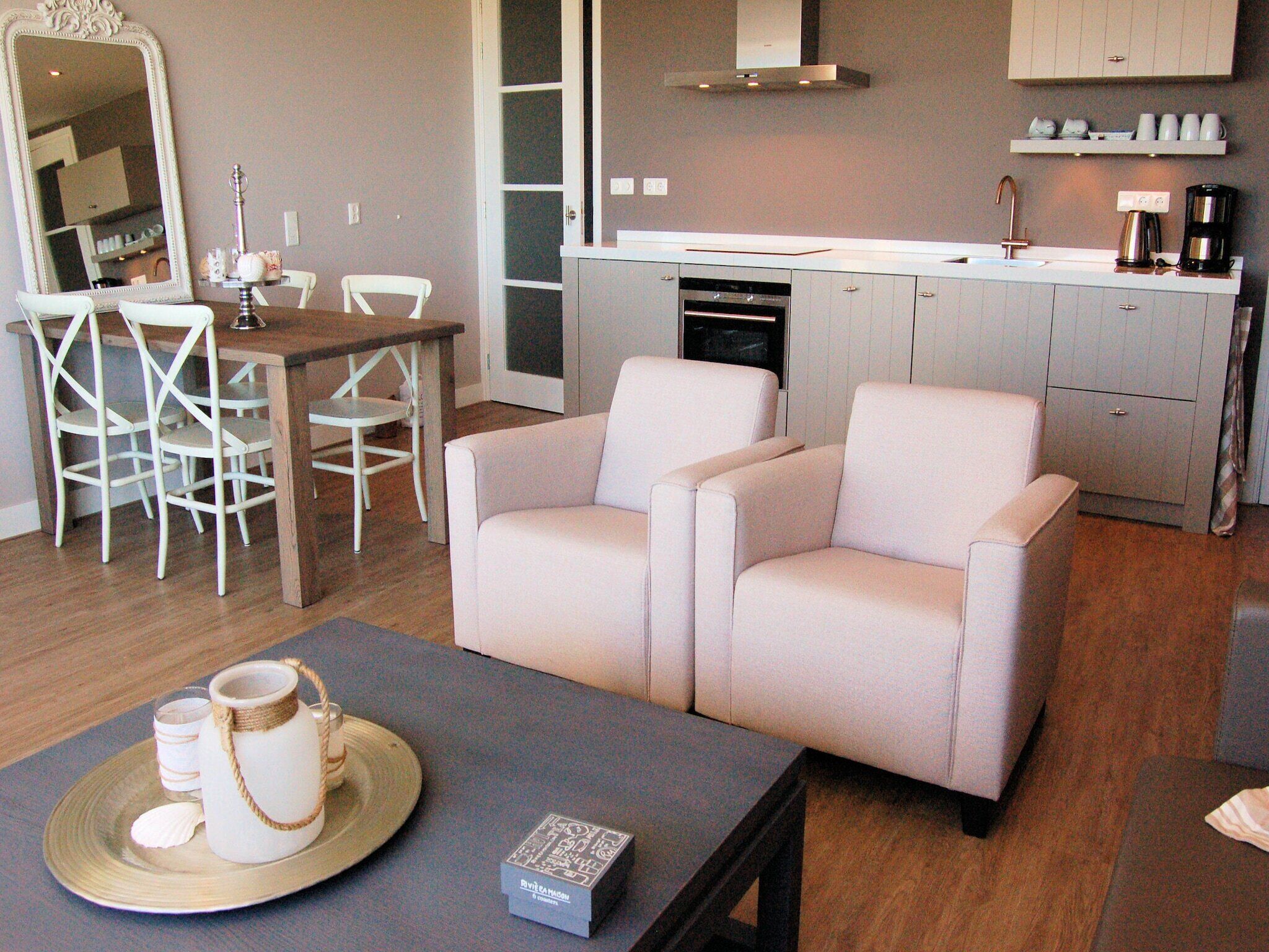 A modern living room with a sofa, a dining table with chairs, a kitchen area with cabinets and appliances, and a large mirror.