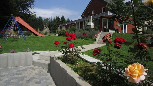 Um imóvel residencial com um escorrega vermelho, um jardim com rosas vermelhas e amarelas, e um caminho de tijolos que leva à entrada.